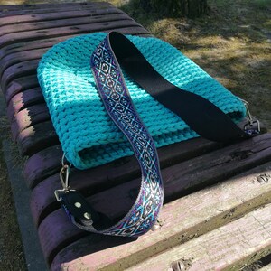 K&ouml;nnte beinhalten: Eine t&uuml;rkisfarbene H&auml;keltasche mit einem schwarzen Riemen und einem dekorativen Schulterriemen mit einem geometrischen Muster in Blau, Lila und Wei&szlig;. Die Tasche liegt auf einer Holzbank.