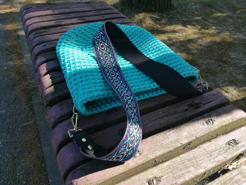 K&ouml;nnte beinhalten: Eine t&uuml;rkisfarbene H&auml;keltasche mit einem schwarzen Riemen und einem dekorativen Schulterriemen mit einem geometrischen Muster in Blau, Lila und Wei&szlig;. Die Tasche liegt auf einer Holzbank.