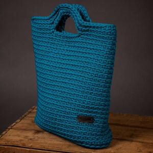 Pode incluir: Uma sacola tote de croch&ecirc; azul-turquesa feita &agrave; m&atilde;o, com formato retangular e duas al&ccedil;as. A sacola tem uma pequena etiqueta retangular de couro presa na frente. A sacola est&aacute; em uma caixa de madeira.