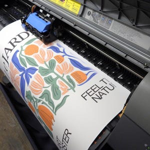 May include: A large format printer producing a white poster with a floral illustration in shades of orange, green, and blue. The text "LE JARDIN" is visible on the left, while "FEEL THE NATURE" and "ART POSTER" are printed in black on the right side.