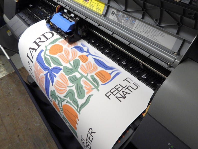 May include: A large format printer producing a white poster with a floral illustration in shades of orange, green, and blue. The text "LE JARDIN" is visible on the left, while "FEEL THE NATURE" and "ART POSTER" are printed in black on the right side.