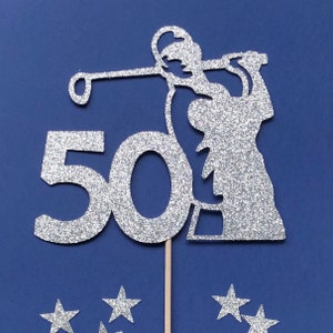 May include: Silver glitter cake topper featuring a golfer in mid-swing and the number 50. The topper is attached to a wooden stick and includes several silver glitter star accents. The background is a solid navy blue.