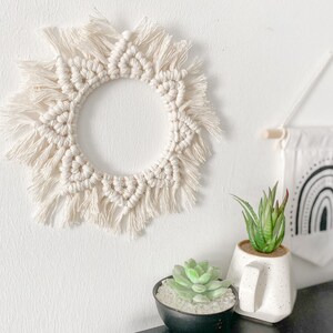 May include: A white macrame wall hanging with a circular shape and fringe details. The hanging is on a white wall with two small potted succulents and a black and white rainbow wall hanging.