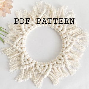 May include: A white macrame wall hanging with a circular shape and fringe details. The text "PDF PATTERN" is printed in black on the wall hanging.