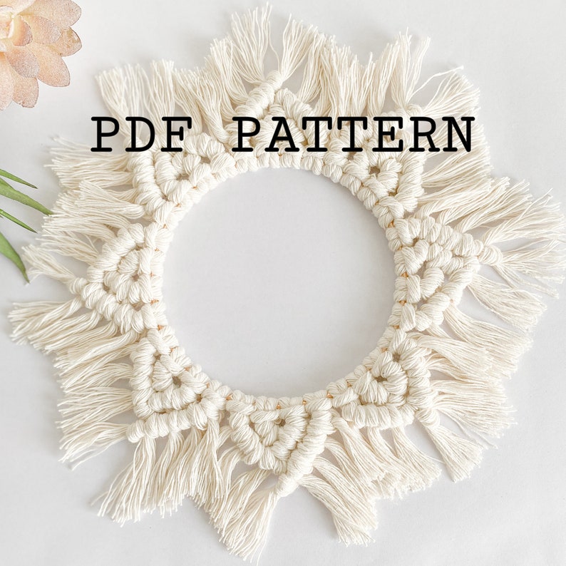 May include: A white macrame wall hanging with a circular shape and fringe details. The text "PDF PATTERN" is printed in black on the wall hanging.