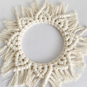 May include: A white macrame wall hanging in the shape of a sun with fringe details.
