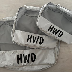 May include: Three light gray packing cubes with mesh tops and zippered closures. Each cube has a white panel with the black letters "HWD". The cubes are different sizes, designed for travel organization.