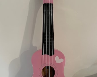 May include: A pink ukulele with white heart designs and the name "ISABELLA" painted on the body. The instrument has black frets, white strings, and tuning pegs. The headstock has the text "CB SKY".