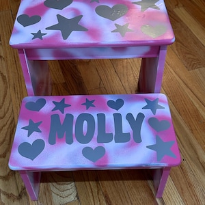 May include: Two-step wooden stool set. The stools are painted pink with a white and pink spray-paint effect. The top of the stools are decorated with gray hearts and stars. The bottom stool has the name "MOLLY" in gray letters.