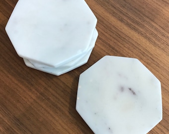 May include: A stack of white marble coasters with an octagonal shape. The coasters have subtle gray veining and are displayed on a wooden surface. The coasters are a decorative item for the home.