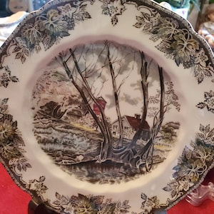 May include: A vintage ceramic plate with a brown and white floral border. The center of the plate features a detailed illustration of a country scene with a farmhouse, trees, and a stream.