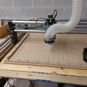 May include: A close-up of a CNC router with a white dust extraction hose attached to the spindle. The router is sitting on a wooden table with a pegboard surface.
