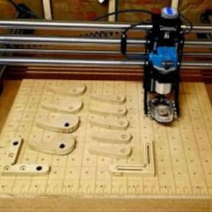 May include: A CNC router machine with a blue router bit attached to the spindle, cutting a pattern into a piece of wood. The wood is on a gridded surface with several other pieces of wood already cut.