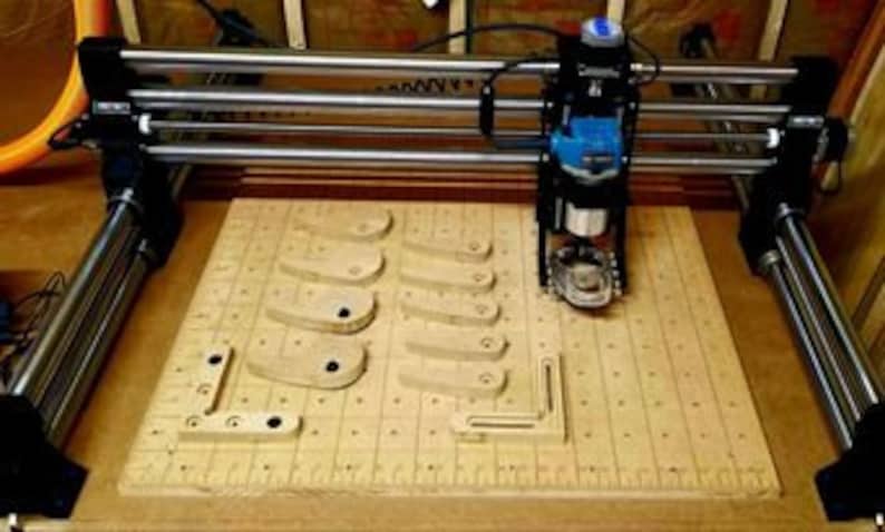 May include: A CNC router machine with a blue router bit attached to the spindle, cutting a pattern into a piece of wood. The wood is on a gridded surface with several other pieces of wood already cut.