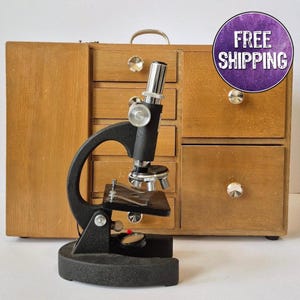May include: A vintage black microscope with silver-colored components stands in front of a wooden cabinet with multiple drawers. The cabinet has a light brown finish and round, silver-colored knobs. The microscope has a circular base and a curved arm.