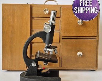 May include: A vintage black microscope with silver-colored components stands in front of a wooden cabinet with multiple drawers. The cabinet has a light brown finish and round, silver-colored knobs. The microscope has a circular base and a curved arm.
