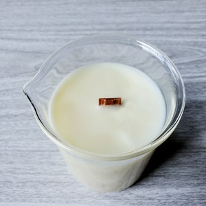 K&ouml;nnte beinhalten: Ein klarer Glashalter mit einer cremewei&szlig;en Kerze und einem Holzdocht. Der Kerzenhalter hat eine einzigartige Form mit einem Ausguss. Die Kerze ist unparf&uuml;miert und hat eine klare, minimalistische &Auml;sthetik.