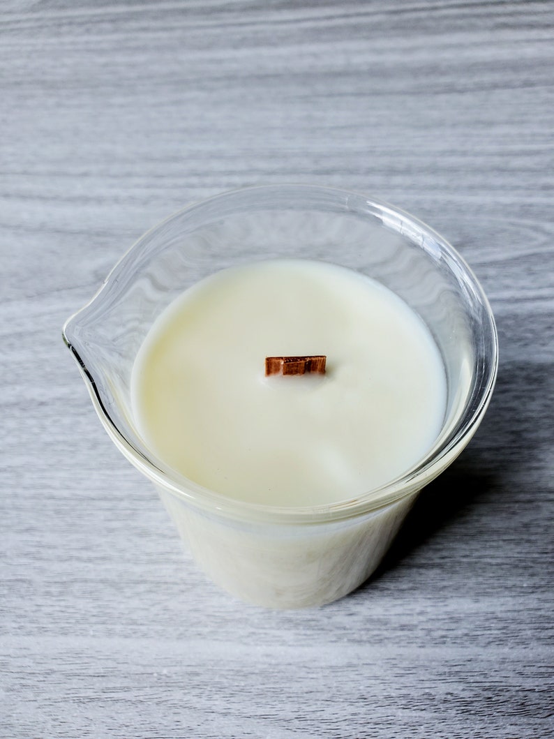 K&ouml;nnte beinhalten: Ein klarer Glashalter mit einer cremewei&szlig;en Kerze und einem Holzdocht. Der Kerzenhalter hat eine einzigartige Form mit einem Ausguss. Die Kerze ist unparf&uuml;miert und hat eine klare, minimalistische &Auml;sthetik.