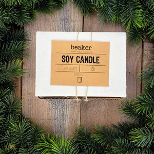 K&ouml;nnte beinhalten: Eine wei&szlig;e Box mit einem braunen Etikett mit der Aufschrift "Soy Candle" und "Clean Burn". Die Box ist mit Bindfaden verschn&uuml;rt und von gr&uuml;nen Kiefernzweigen umgeben, vor einem Holzgrund. Die Kerze ist 250 ml.