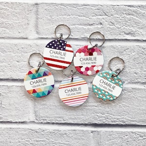 May include: Five colorful round dog tags with silver rings. Each tag has the name "CHARLIE" and a phone number "123.456.7890". The tags feature different patterns: American flag, pink hexagons, rainbow stripes, blue and green geometric shapes, and turquoise with rainbows.