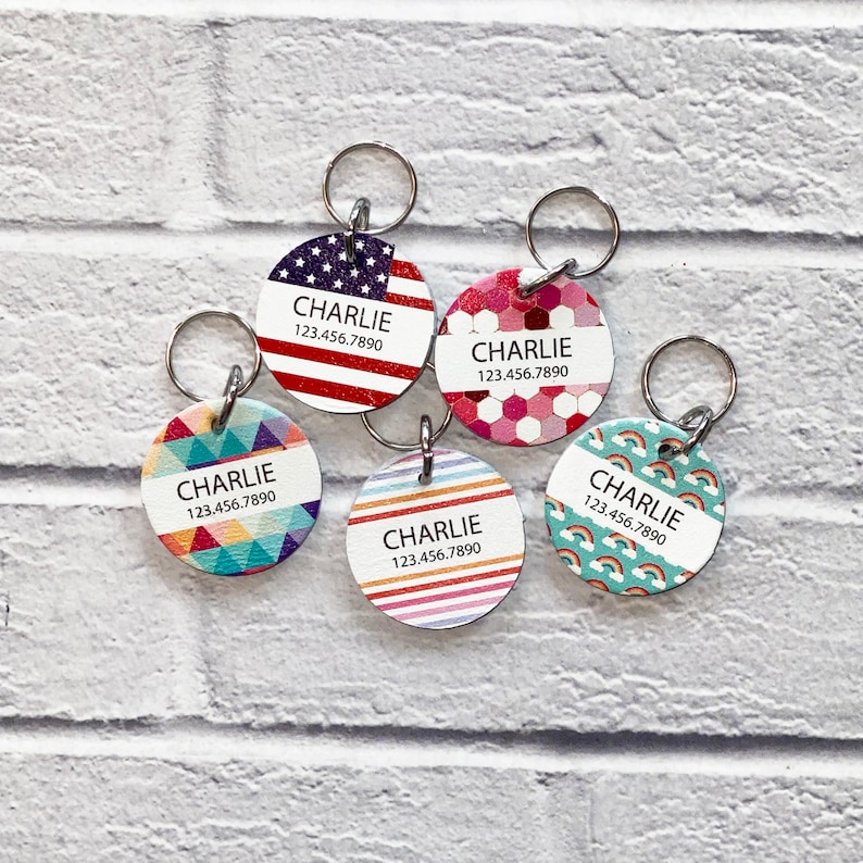 May include: Five colorful round dog tags with silver rings. Each tag has the name "CHARLIE" and a phone number "123.456.7890". The tags feature different patterns: American flag, pink hexagons, rainbow stripes, blue and green geometric shapes, and turquoise with rainbows.