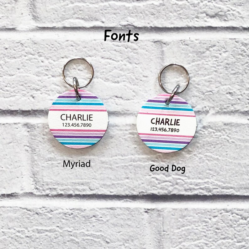 May include: Two round dog tags with a striped design in pink, blue, and purple. The tags have the name "CHARLIE" and the phone number "123.456.7890" printed on them. The tags are labeled "Myriad" and "Good Dog" below them.