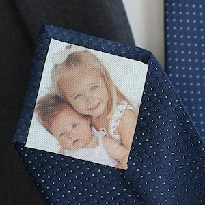 Pode incluir: Uma gravata azul marinho com bolinhas brancas, com um inserto de foto quadrado de duas crianças. A foto mostra uma menina pequena segurando um bebê.