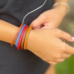 May include: A set of six leather bracelets with engraved text. The bracelets are in various colours, including brown, red, blue, and yellow. The text on the bracelets includes names, dates, and a bible verse.