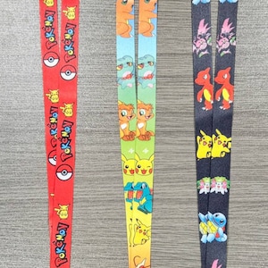 May include: Three colourful Pokemon lanyards featuring different Pokemon characters. The lanyards are red, yellow and black with a variety of Pokemon characters on them. The lanyards have a metal clip at the end.
