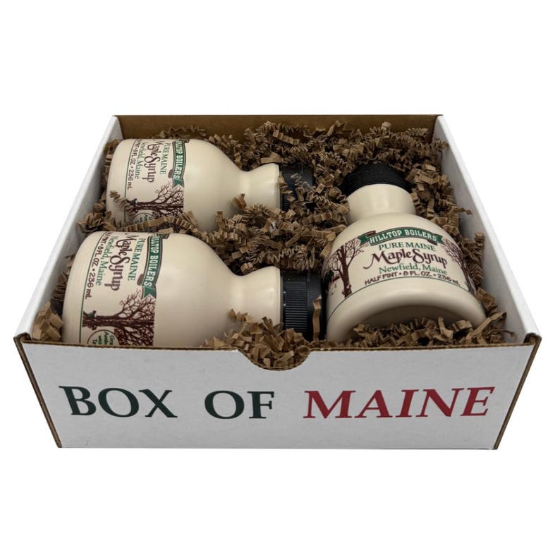 May include: A gift box labeled "BOX OF MAINE" contains three bottles of maple syrup. The bottles are off-white with black caps and labels that read "Pure Maine Maple Syrup." The box is filled with brown packing material.