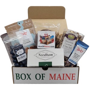 May include: A white box with the words "BOX OF MAINE" printed in green on the front. The box is open and filled with various Maine-made treats, including chocolate caramels, chocolate sea salt cashews, blueberry sea salt cashews, and a box of Maine Needham chocolates.