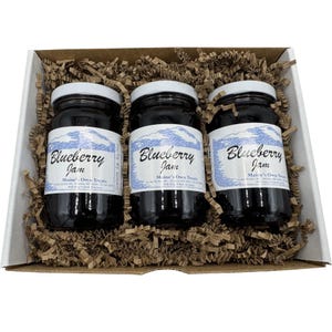 May include: A gift box containing three jars of blueberry jam. Each jar has a white lid and a label with the words "Blueberry Jam" and "Maine's Own Treats". The jars are nestled in brown paper filler.