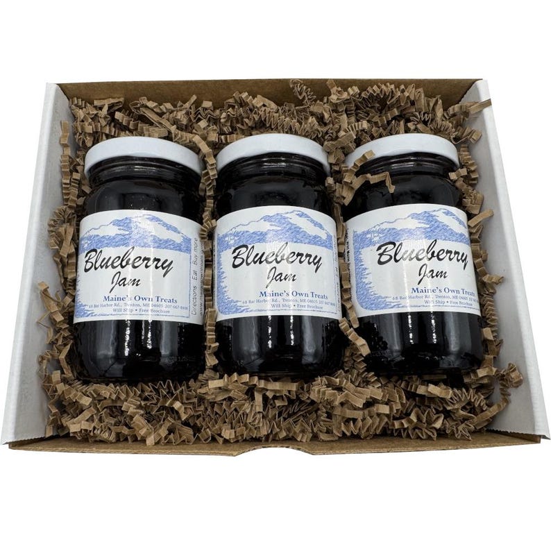 May include: A gift box containing three jars of blueberry jam. Each jar has a white lid and a label with the words "Blueberry Jam" and "Maine's Own Treats". The jars are nestled in brown paper filler.