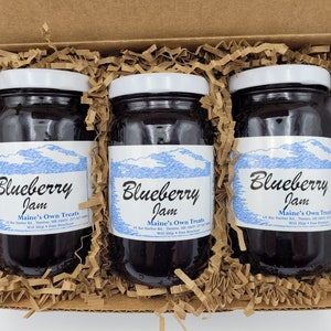 May include: Three glass jars of blueberry jam in a brown cardboard box with shredded paper. The jars have white lids and blue and white labels that say "Blueberry Jam" and "Maine's Own Treats".