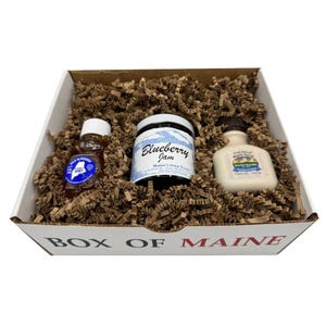 May include: A gift box filled with brown packing material, containing three food items. There is a small bottle of honey, a jar of blueberry jam, and a bottle of maple syrup. The box is white with the words "BOX OF MAINE" printed on the front.