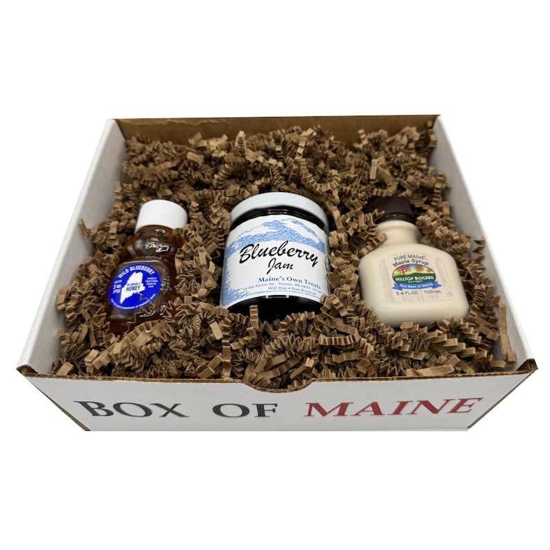 May include: A gift box filled with brown packing material, containing three food items. There is a small bottle of honey, a jar of blueberry jam, and a bottle of maple syrup. The box is white with the words "BOX OF MAINE" printed on the front.