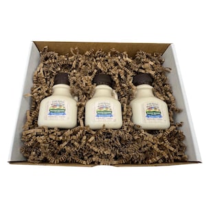 May include: A gift box containing three small, white maple syrup bottles with dark brown caps. Each bottle has a label that reads "Pure Maine Maple Syrup" and "Hilltop Boilers". The bottles are nestled in brown paper filler.