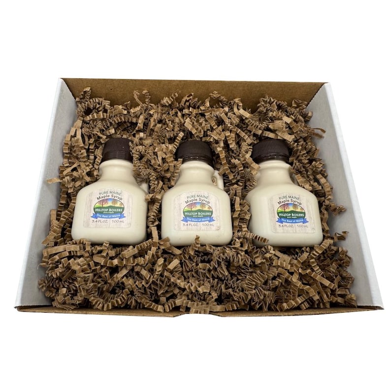 May include: A gift box containing three small, white maple syrup bottles with dark brown caps. Each bottle has a label that reads "Pure Maine Maple Syrup" and "Hilltop Boilers". The bottles are nestled in brown paper filler.