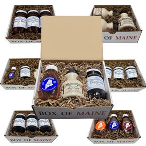 May include: Assortment of Maine food products in gift boxes. Jars of strawberry and blueberry jam, maple syrup, and honey are visible. The boxes are filled with crinkle paper and labeled "Box of Maine."