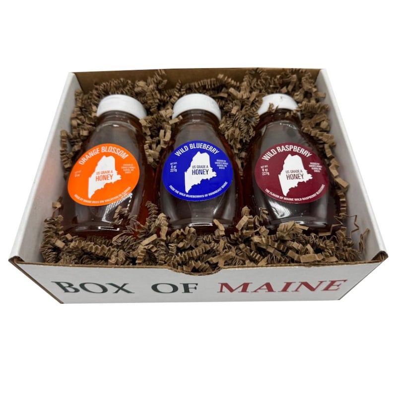 May include: A gift box labeled "Box of Maine" contains three glass bottles of honey. Each bottle has a white cap and a colorful label: orange for Orange Blossom, blue for Wild Blueberry, and red for Wild Raspberry. The bottles are nestled in brown packing material.