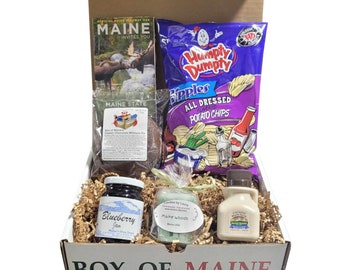May include: A brown cardboard box with the words "Box of Maine" printed on the front. The box is filled with various Maine-themed products, including a map, potato chips, a chocolate pie, blueberry jam, a candle, and maple syrup.
