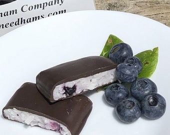 Puede incluir: Dos golosinas cubiertas de chocolate con un relleno blanco y arándanos. Las golosinas están en un plato blanco con una hoja verde y arándanos.