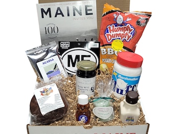 May include: A brown cardboard box with the words "Box of Maine" printed on the front. The box is filled with various Maine-themed items, including a magazine, snacks, a jar of jam, a bottle of maple syrup, and a candle.