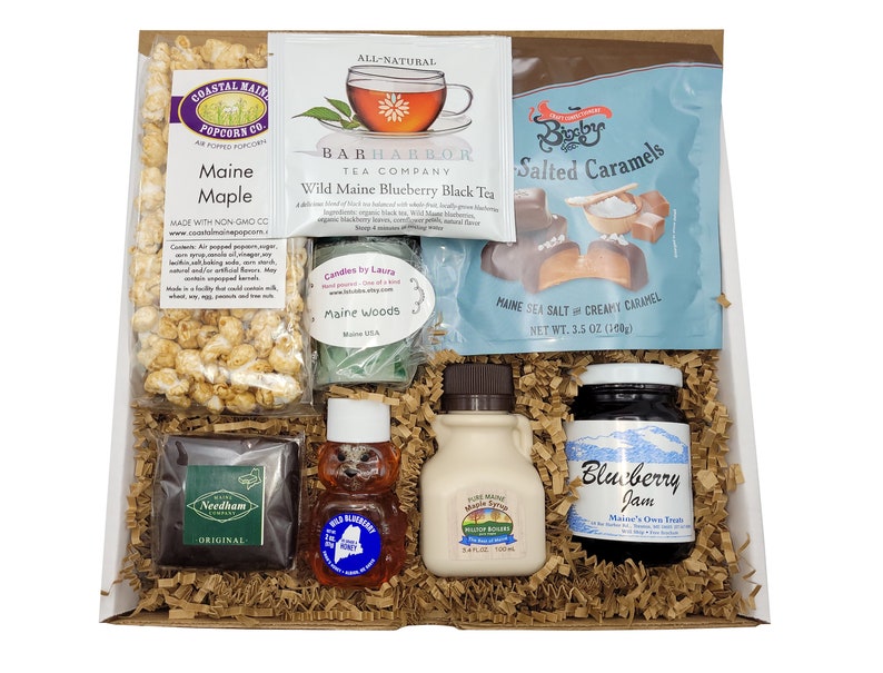 May include: A gift basket filled with Maine-themed treats, including popcorn, tea, caramels, honey, maple syrup, and blueberry jam. The basket is lined with brown paper shred.