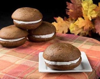Puede incluir: Cuatro pasteles whoopie de chocolate con glaseado blanco sobre un mantel a cuadros. Los pasteles whoopie están apilados uno encima del otro.