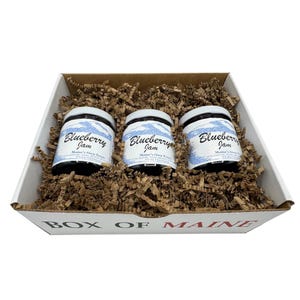 May include: A gift box containing three jars of blueberry jam. The jars have white labels with blue text that reads "Blueberry Jam" and "Maine's Own Treats." The jars are nestled in brown paper filler inside a white box with "BOX OF MAINE" printed on the front.