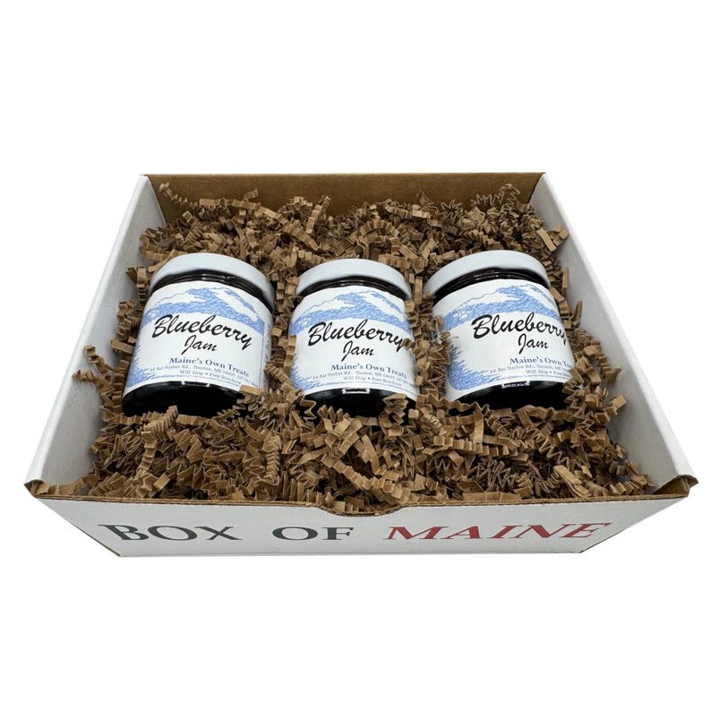 May include: A gift box containing three jars of blueberry jam. The jars have white labels with blue text that reads "Blueberry Jam" and "Maine's Own Treats." The jars are nestled in brown paper filler inside a white box with "BOX OF MAINE" printed on the front.