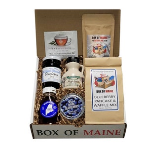 May include: A brown cardboard box with the words "Box of Maine" printed on the front. The box is filled with various Maine-themed food items, including blueberry jam, maple syrup, wild blueberry tea, wild blueberries, and a blueberry pancake and waffle mix.