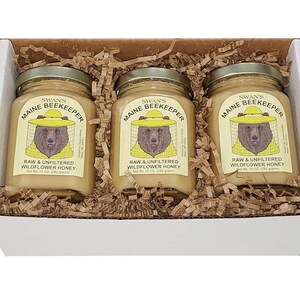 May include: A white box contains three jars of honey. Each jar has a label with a bear wearing a beekeeper hat. The labels read "Maine Beekeeper" and "Raw & Unfiltered Wildflower Honey". Each jar contains 10 oz (283 grams) of honey.