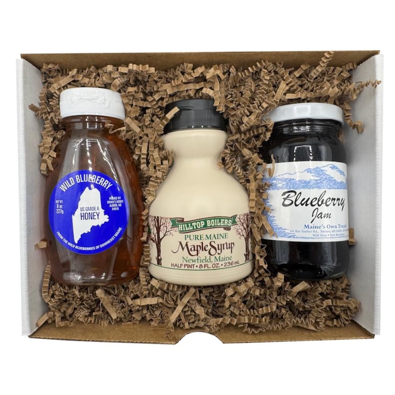 May include: A gift box containing three food items: a bottle of Wild Blueberry Honey, a bottle of Pure Maine Maple Syrup, and a jar of Blueberry Jam. The honey bottle is clear, the syrup bottle is cream-colored, and the jam jar is black.